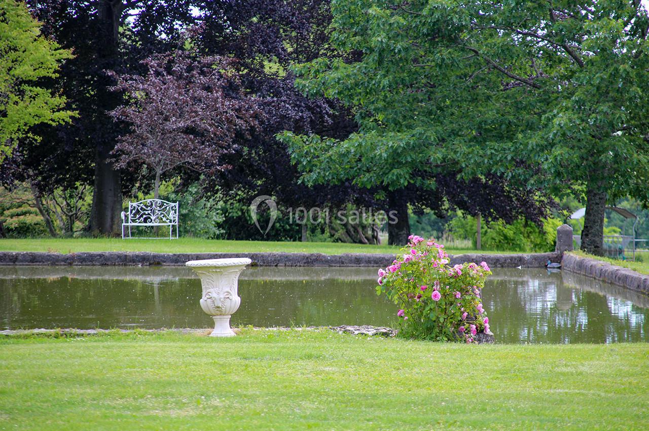 Banc blanc et vase décoratif près d'un étang entouré d'arbres et de fleurs dans un jardin verdoyant.