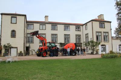 Un groupe de personnes pose devant un tracteur rouge et une machine agricole, devant un grand bâtiment ancien.