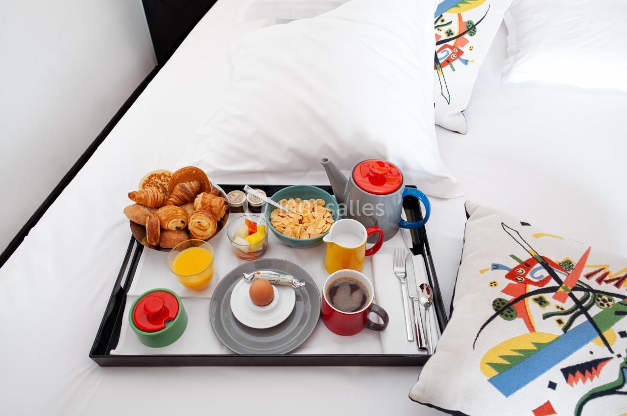 Plateau de petit-déjeuner avec viennoiseries, œuf, céréales, boissons chaudes et jus, posé sur un lit blanc.