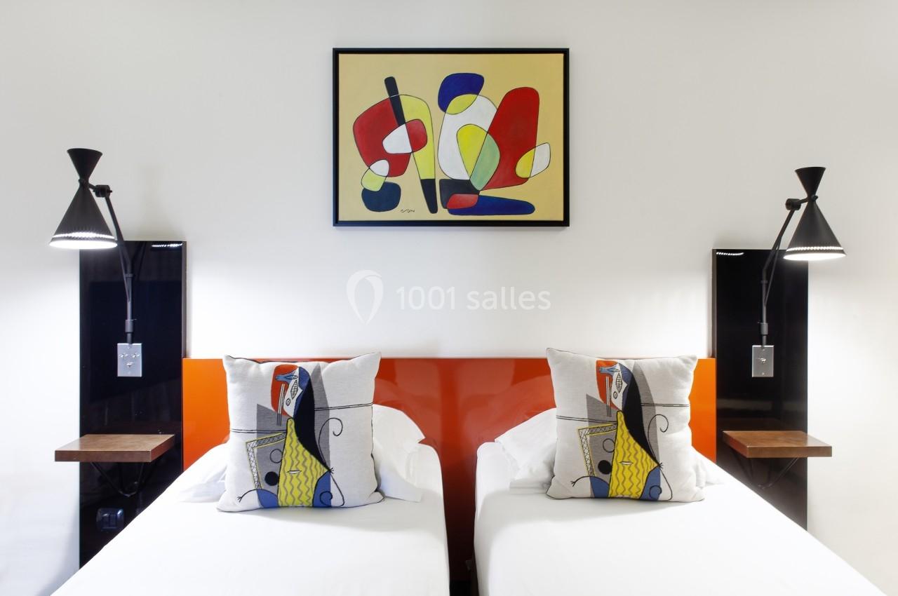 Deux lits simples avec des coussins décoratifs, éclairés par des lampes murales, sous un tableau coloré abstrait.