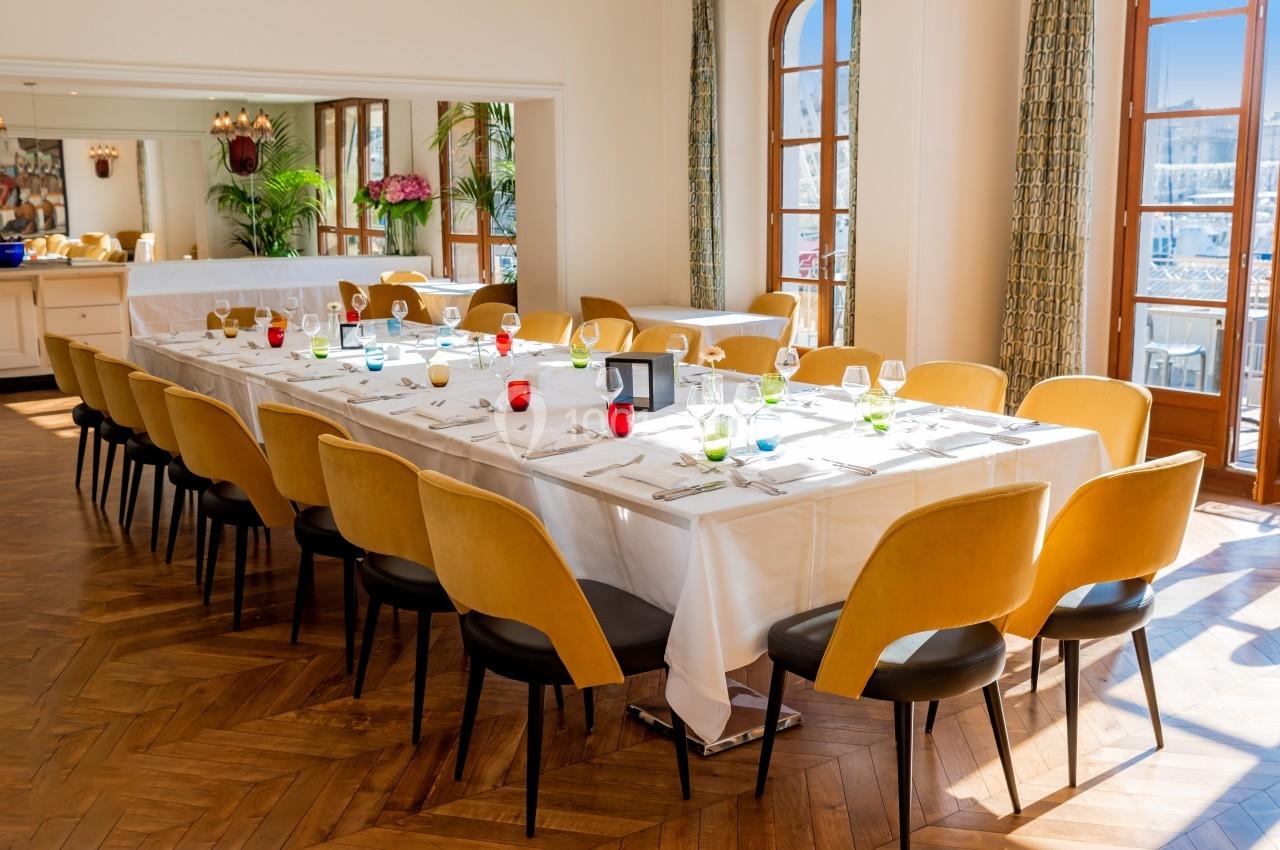 Salle lumineuse avec une grande table dressée, chaises jaunes, vaisselle colorée et grandes fenêtres donnant sur l'extérieur.