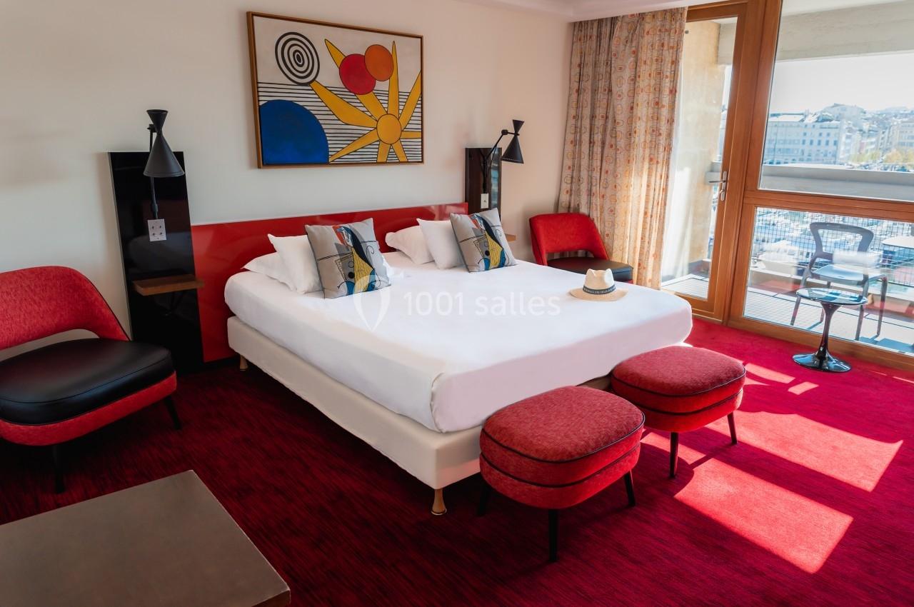 Chambre d'hôtel lumineuse avec lit double, fauteuils rouges, tableau coloré et balcon donnant sur une vue urbaine.