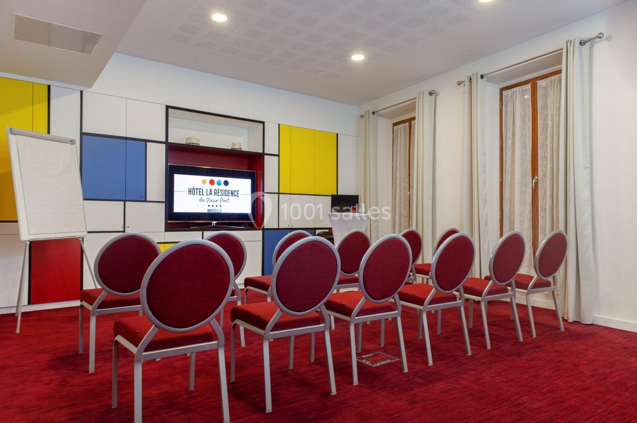 Salle de réunion avec chaises rouges alignées, écran allumé, paperboard et murs colorés.