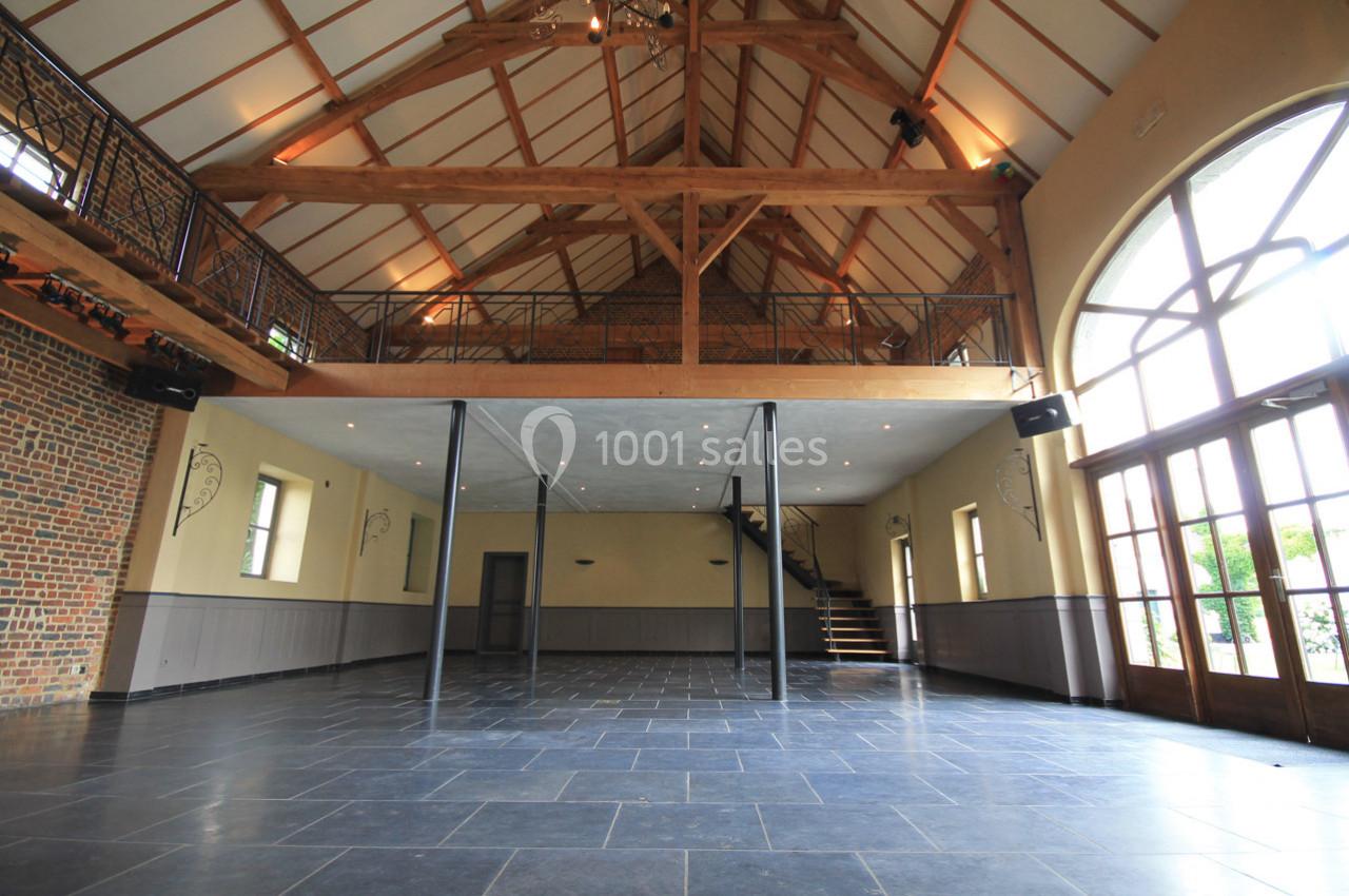 Salle spacieuse avec charpente en bois apparente, sol en pierre sombre, mezzanine et grande baie vitrée.