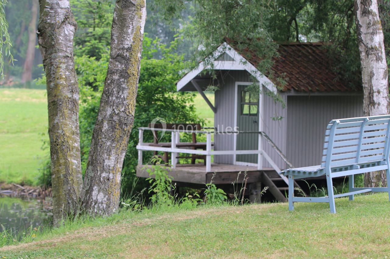 Petite cabane en bois avec terrasse au bord d'un étang, entourée d'arbres et d'une pelouse avec un banc.