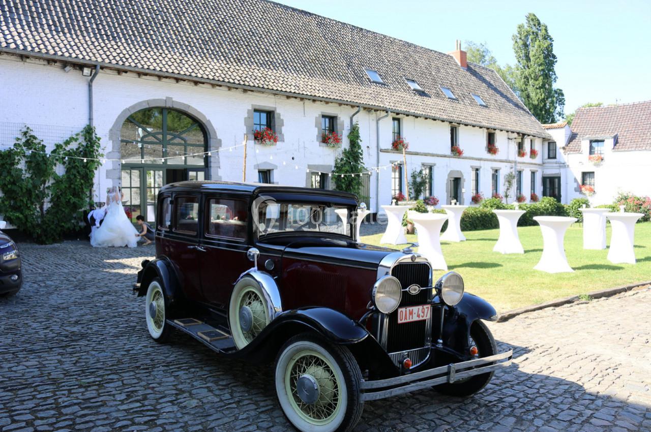 Voiture ancienne noire et bordeaux stationnée sur des pavés devant un bâtiment blanc avec des fenêtres et des plantes.