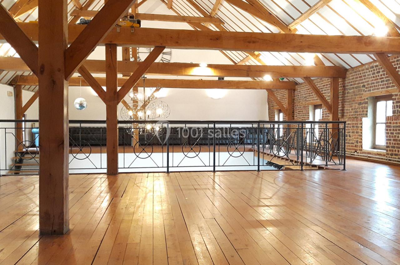 Plancher en bois clair sous une charpente apparente, avec une rambarde métallique et des murs en briques.