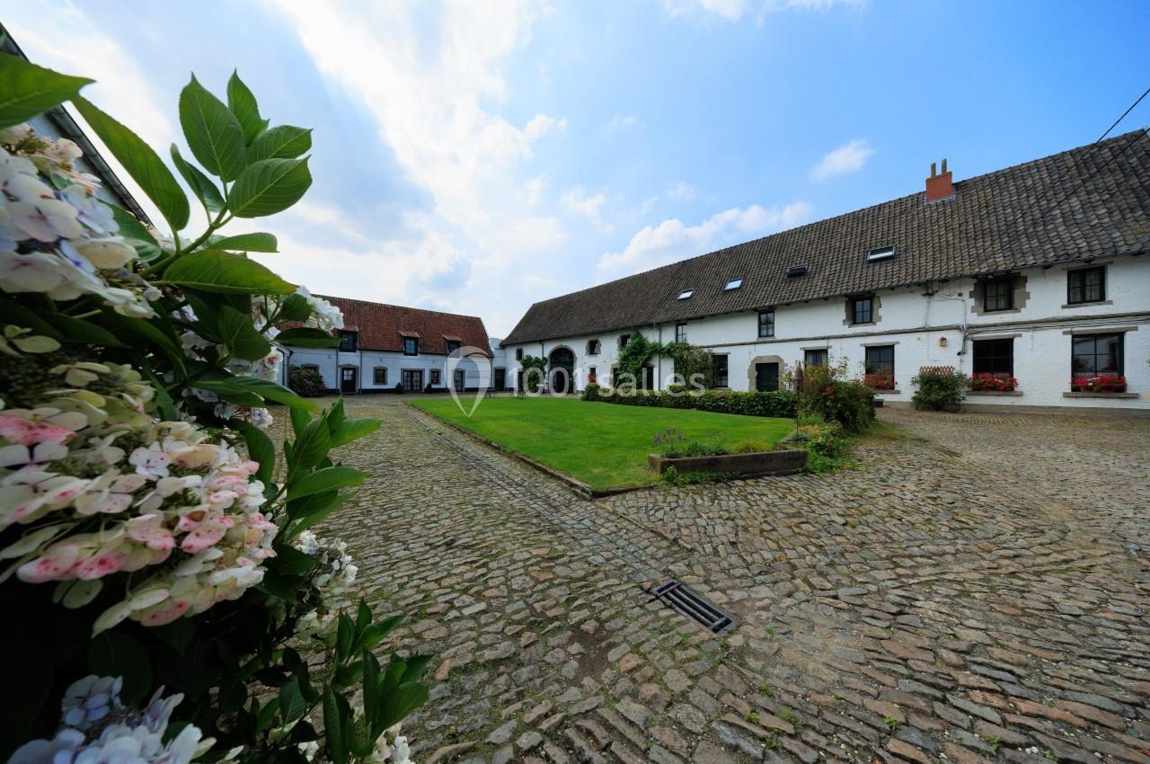 Cour pavée entourée de bâtiments blancs à toit en tuiles, avec des fleurs au premier plan et un ciel partiellement nuageux.