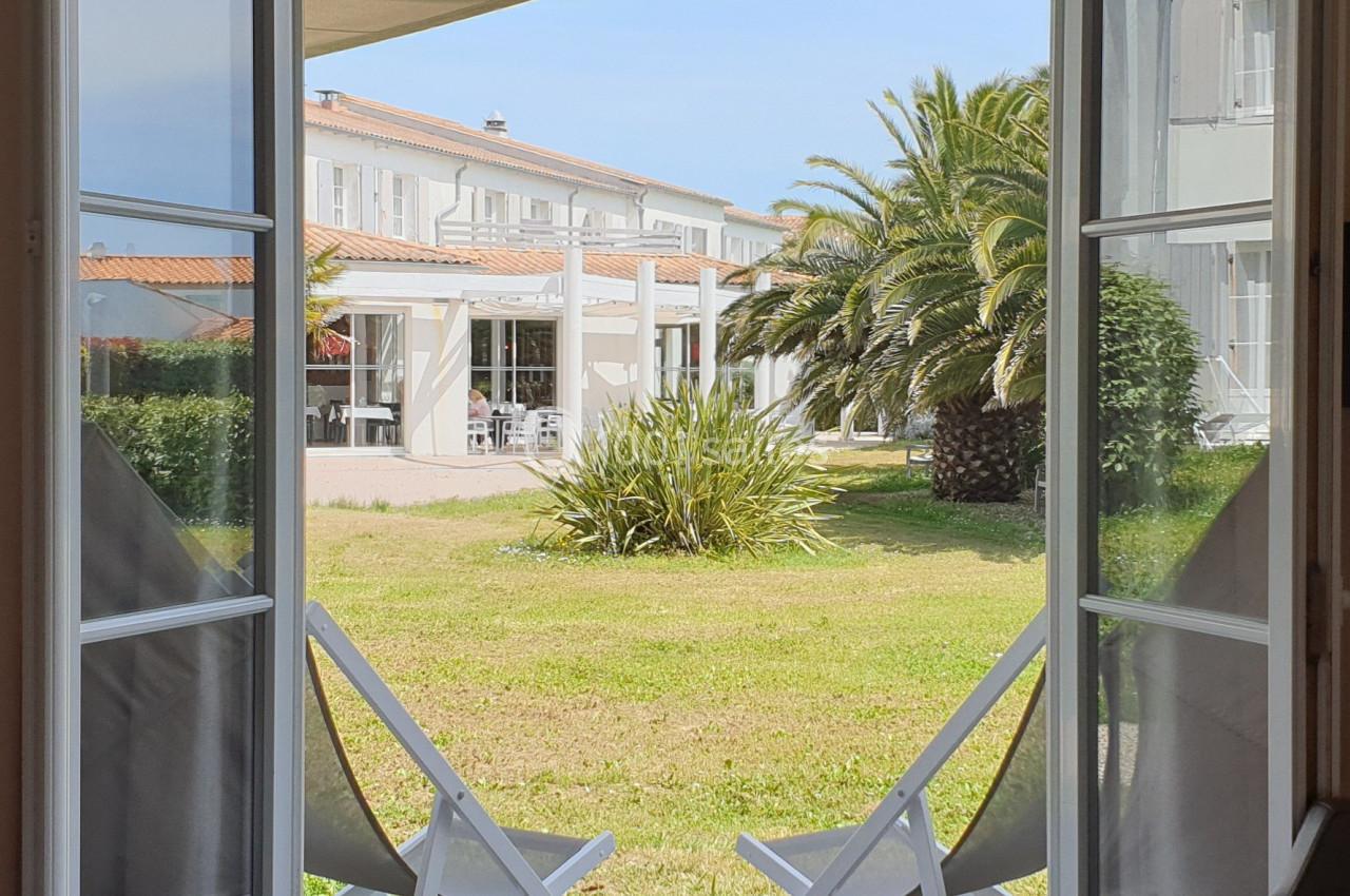 Vue depuis une porte-fenêtre ouverte sur une terrasse en bois, deux chaises et un jardin avec palmiers et bâtiment blanc.