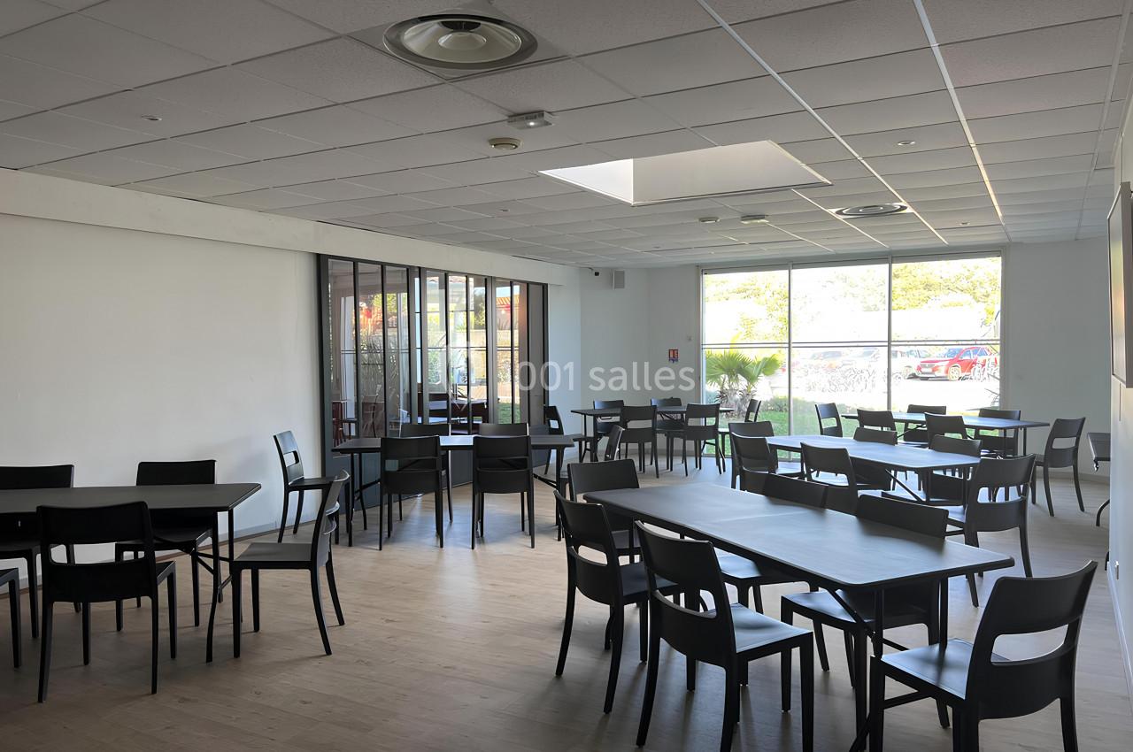 Salle lumineuse avec tables et chaises noires, grandes fenêtres laissant entrer la lumière naturelle.