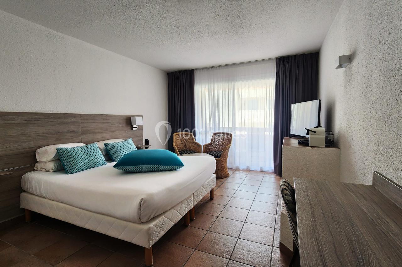 Chambre lumineuse avec lit double, bureau, fauteuils en osier, télévision et grande fenêtre avec rideaux.