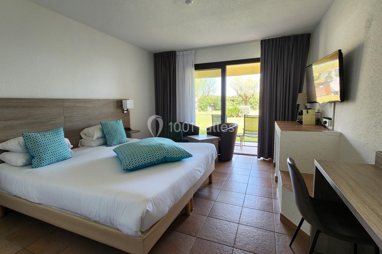 Chambre lumineuse avec lit double, bureau, fauteuils, et baie vitrée donnant sur un jardin.