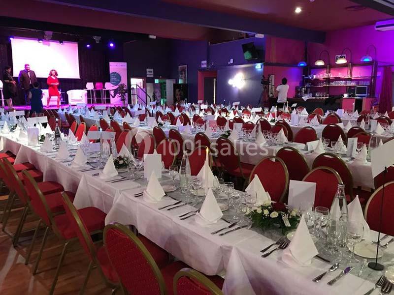Salle de réception avec de longues tables dressées, nappes blanches, serviettes pliées et décorations florales.