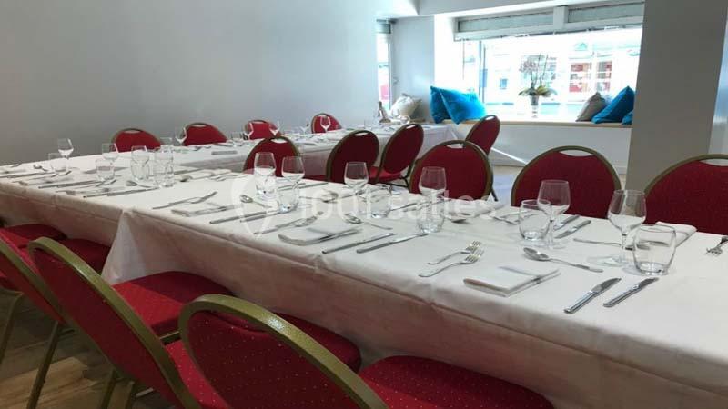 Salle avec longues tables dressées, nappes blanches, chaises rouges et lumière naturelle provenant de grandes fenêtres.