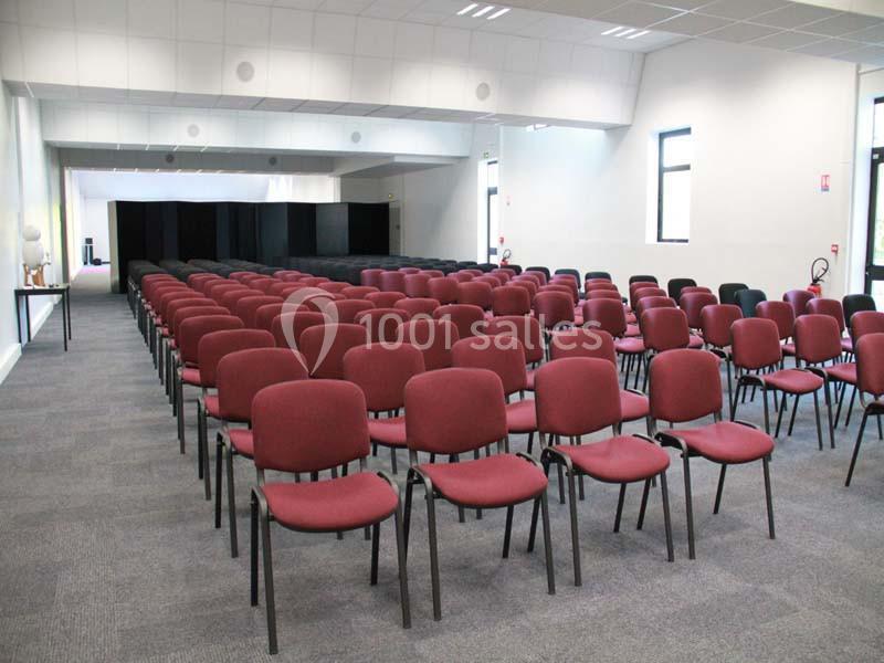 Salle de conférence avec des rangées de chaises rouges alignées sur un sol en moquette grise.