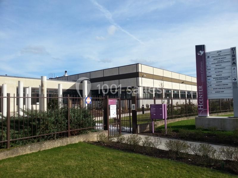 Bâtiment moderne entouré d'une clôture, avec un panneau d'information et un espace vert au premier plan.