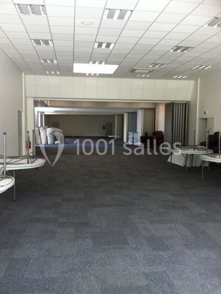 Salle vide avec moquette grise, tables pliées sur les côtés et plafond équipé de luminaires encastrés.