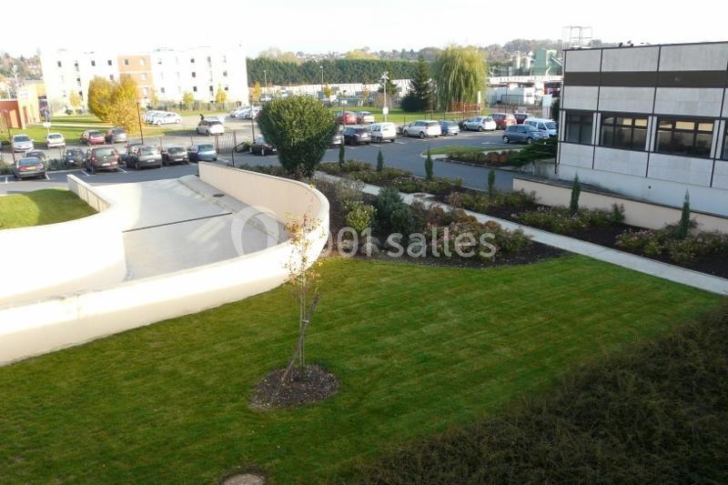 Vue d'un espace extérieur avec pelouse, arbustes, allée et bâtiments entourés de parkings en arrière-plan.