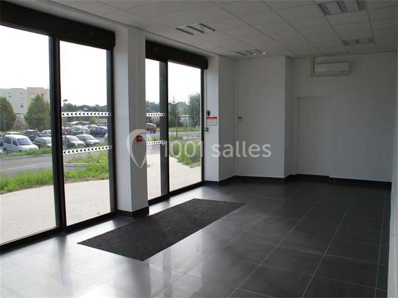 Intérieur d'un espace vide avec sol carrelé, grandes baies vitrées donnant sur un parking et un extérieur verdoyant.