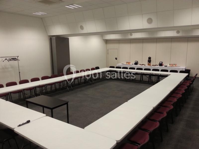 Salle de réunion vide avec des tables disposées en U, des chaises rouges et un portant à vêtements dans un coin.