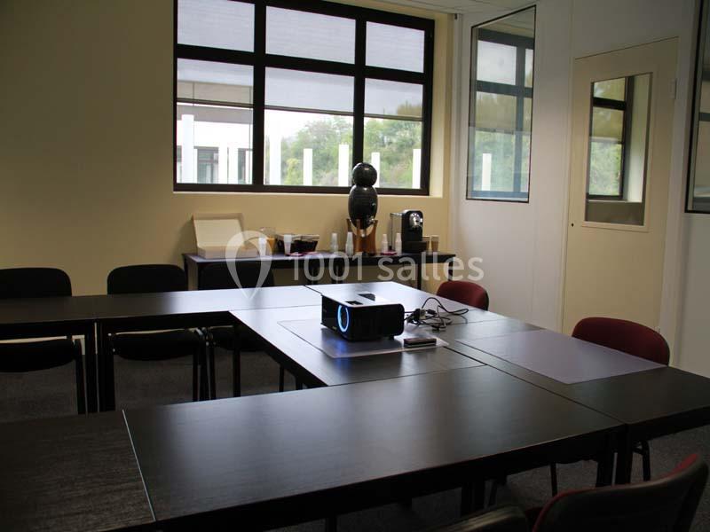 Salle de réunion avec tables en U, vidéoprojecteur allumé au centre et coin café en arrière-plan.