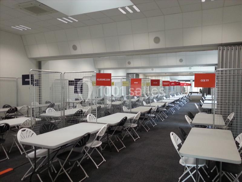 Salle avec des tables et des chaises alignées, séparées par des grilles et des panneaux suspendus indiquant des sections.