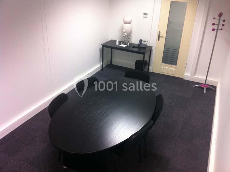 Salle de réunion avec une table ovale noire, six chaises, un bureau avec téléphone et une porte en arrière-plan.