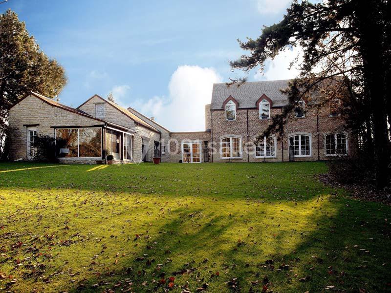Grande maison en pierre avec des fenêtres cintrées, entourée d'une pelouse et d'arbres sous un ciel dégagé.