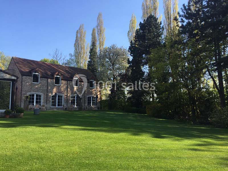 Maison en briques avec toit en tuiles, entourée d'un grand jardin verdoyant et d'arbres sous un ciel dégagé.
