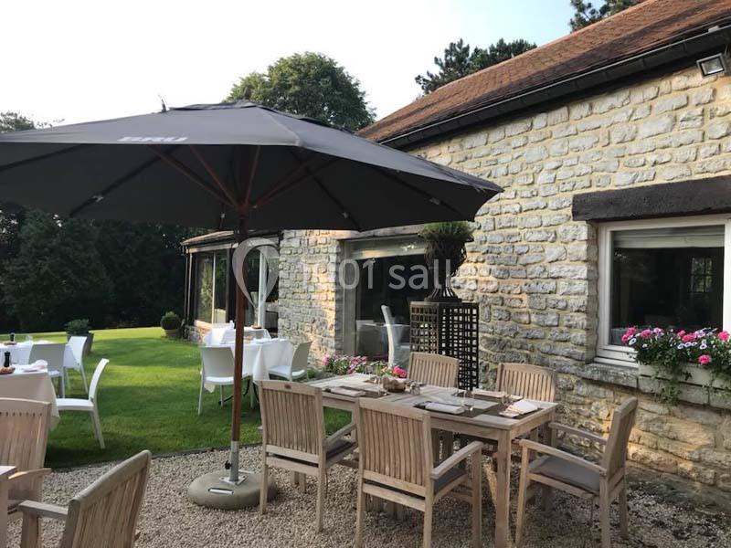 Terrasse extérieure avec tables en bois, chaises blanches et parasol, devant un bâtiment en pierre entouré de verdure.