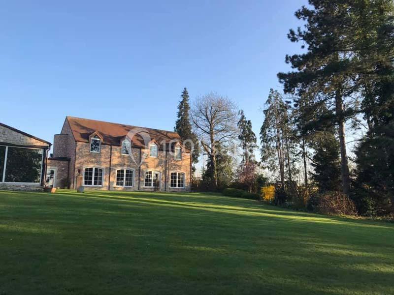 Grande maison en briques avec toit en pente, entourée d'arbres et donnant sur une pelouse verdoyante par temps clair.