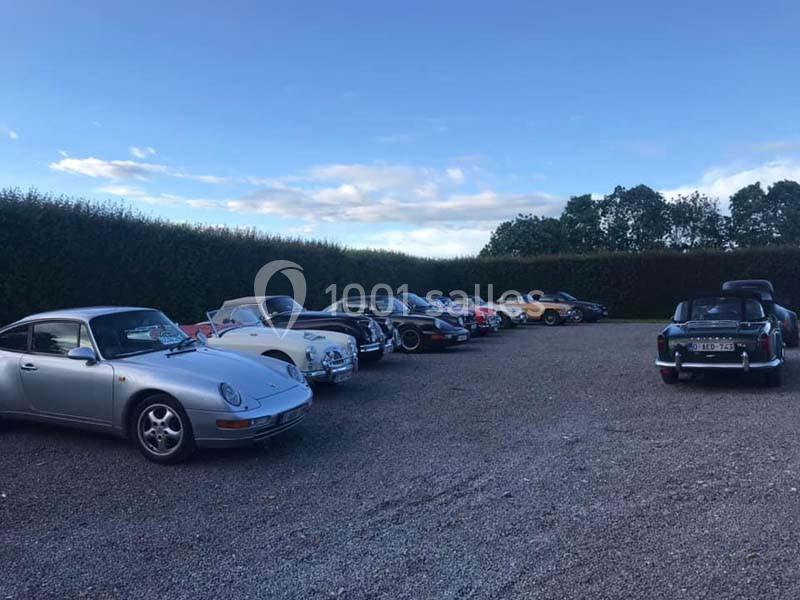 Rangée de voitures anciennes et classiques stationnées sur un parking en gravier, entouré de haies et d'arbres.