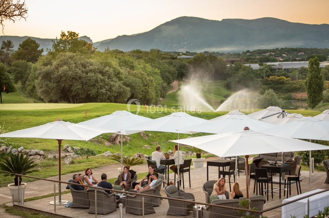 Terrasse avec des tables et des parasols, vue sur un terrain de golf verdoyant et des montagnes en arrière-plan.