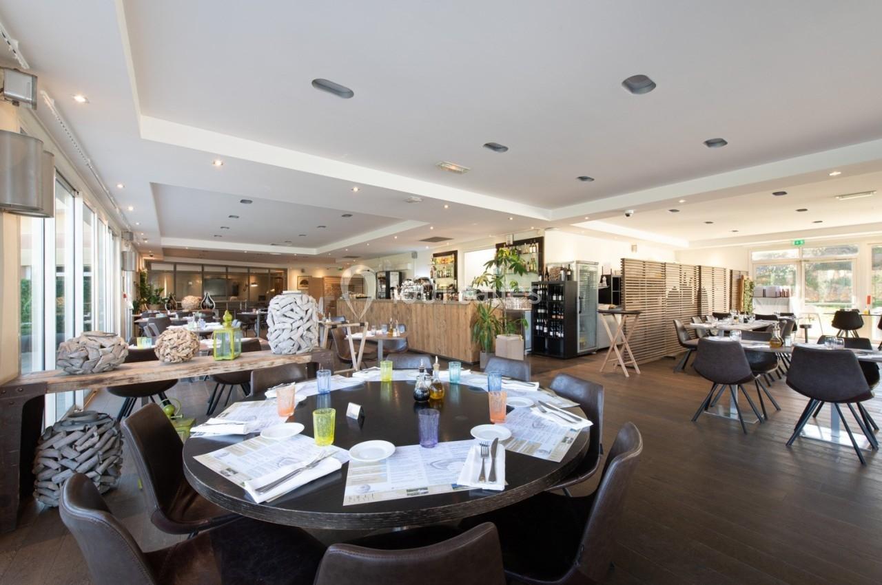 Salle de restaurant lumineuse avec tables dressées, chaises noires et grandes baies vitrées donnant sur l'extérieur.