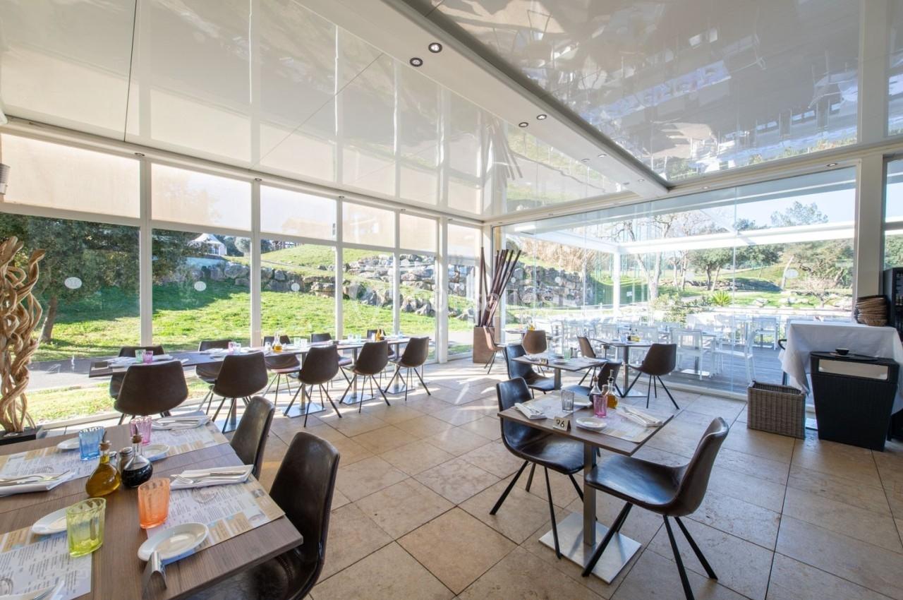Salle de restaurant lumineuse avec de grandes baies vitrées donnant sur un jardin verdoyant.