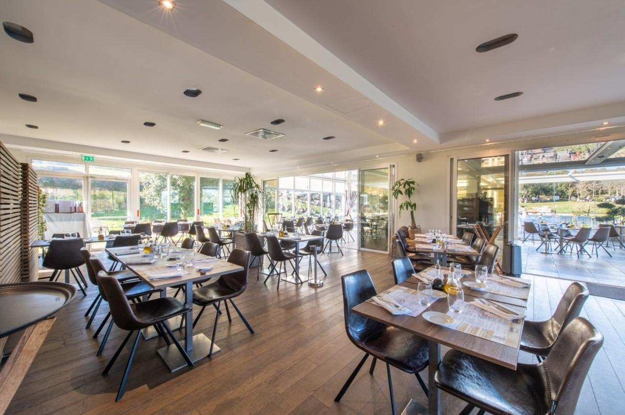 Salle de restaurant lumineuse avec tables dressées, chaises modernes et grandes baies vitrées donnant sur l'extérieur.