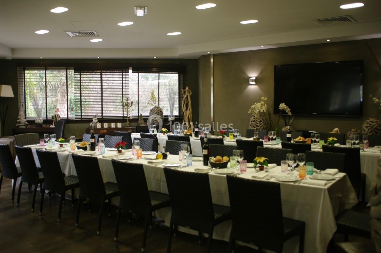 Salle de réception avec de longues tables dressées, chaises noires, décoration élégante et grande télévision murale.