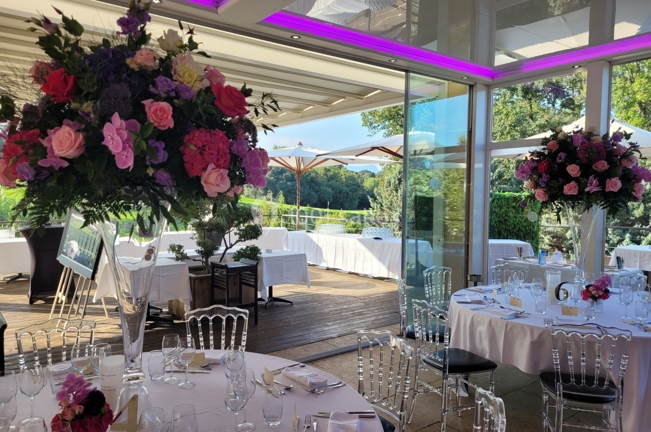Salle de réception lumineuse avec tables dressées, grandes compositions florales et vue sur une terrasse arborée.