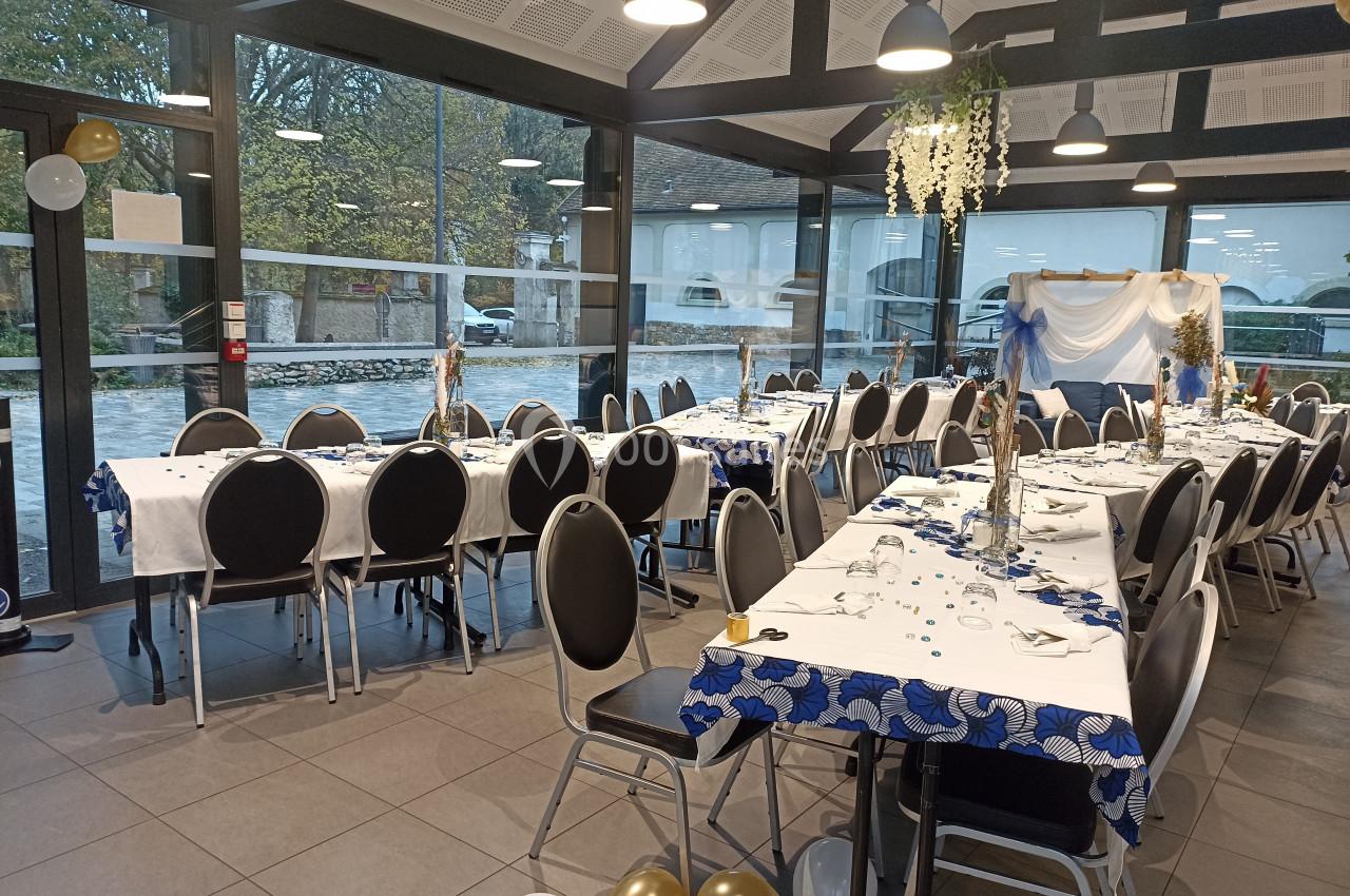 Salle décorée pour un événement avec tables dressées, nappes blanches et bleues, et vue sur l'extérieur vitré.