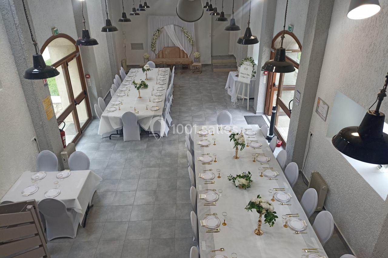 Salle de réception lumineuse avec deux grandes tables dressées, décorées de feuillages et de vaisselle blanche.