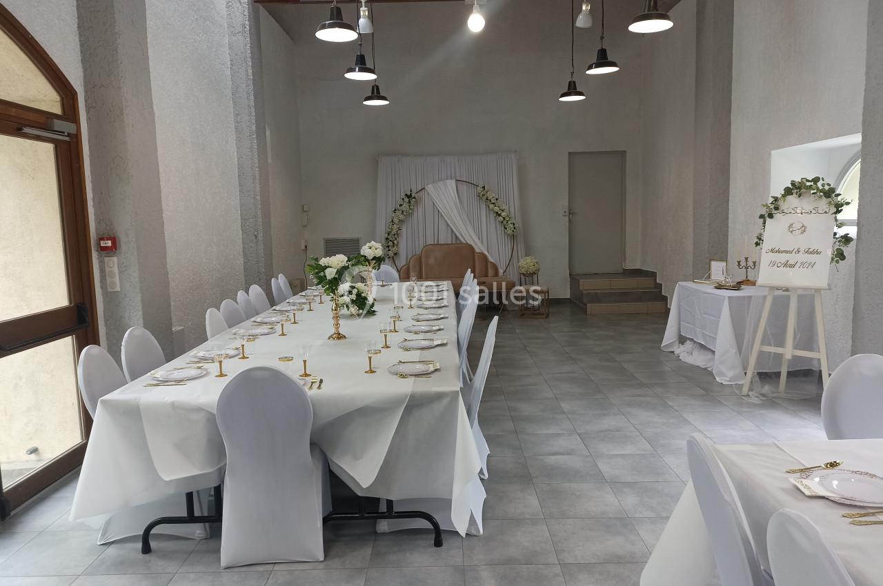 Salle décorée pour un événement avec une grande table dressée, des chaises blanches et un espace d'arche florale.