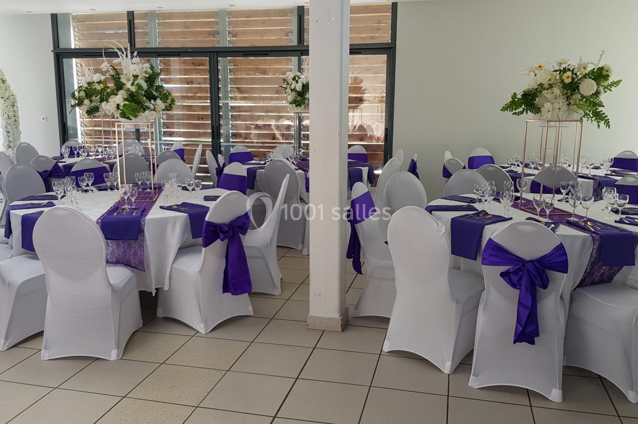 Salle décorée pour un événement avec des tables rondes dressées, nappes blanches et chemins de table violets.