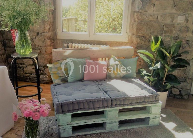 Banc en palettes avec coussins colorés, entouré de plantes et fleurs, dans une pièce lumineuse aux murs en pierre.