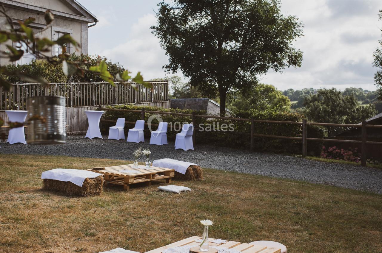 Espace extérieur avec tables en palettes, bottes de paille couvertes de tissu blanc et chaises alignées près d'une maison.
