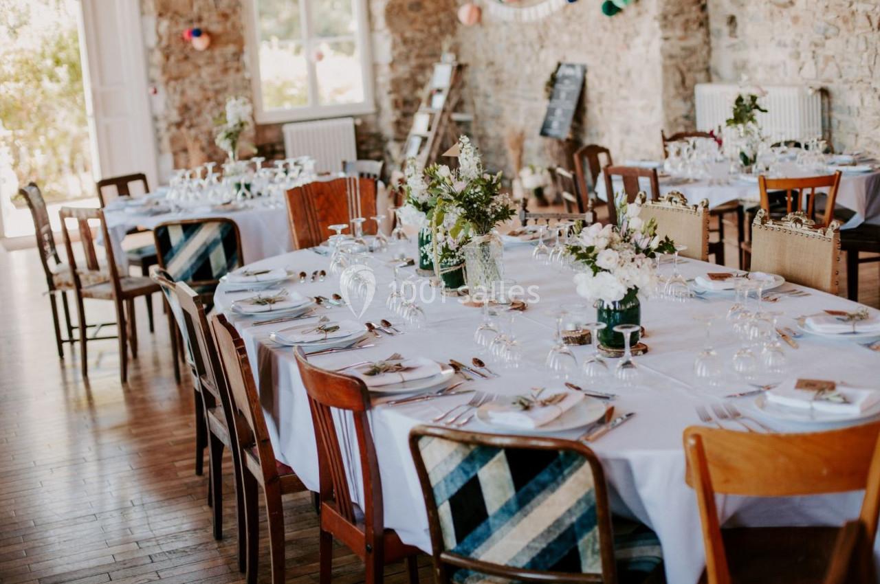 Tables de réception décorées avec nappes blanches, fleurs et vaisselle, dans une salle aux murs en pierre.