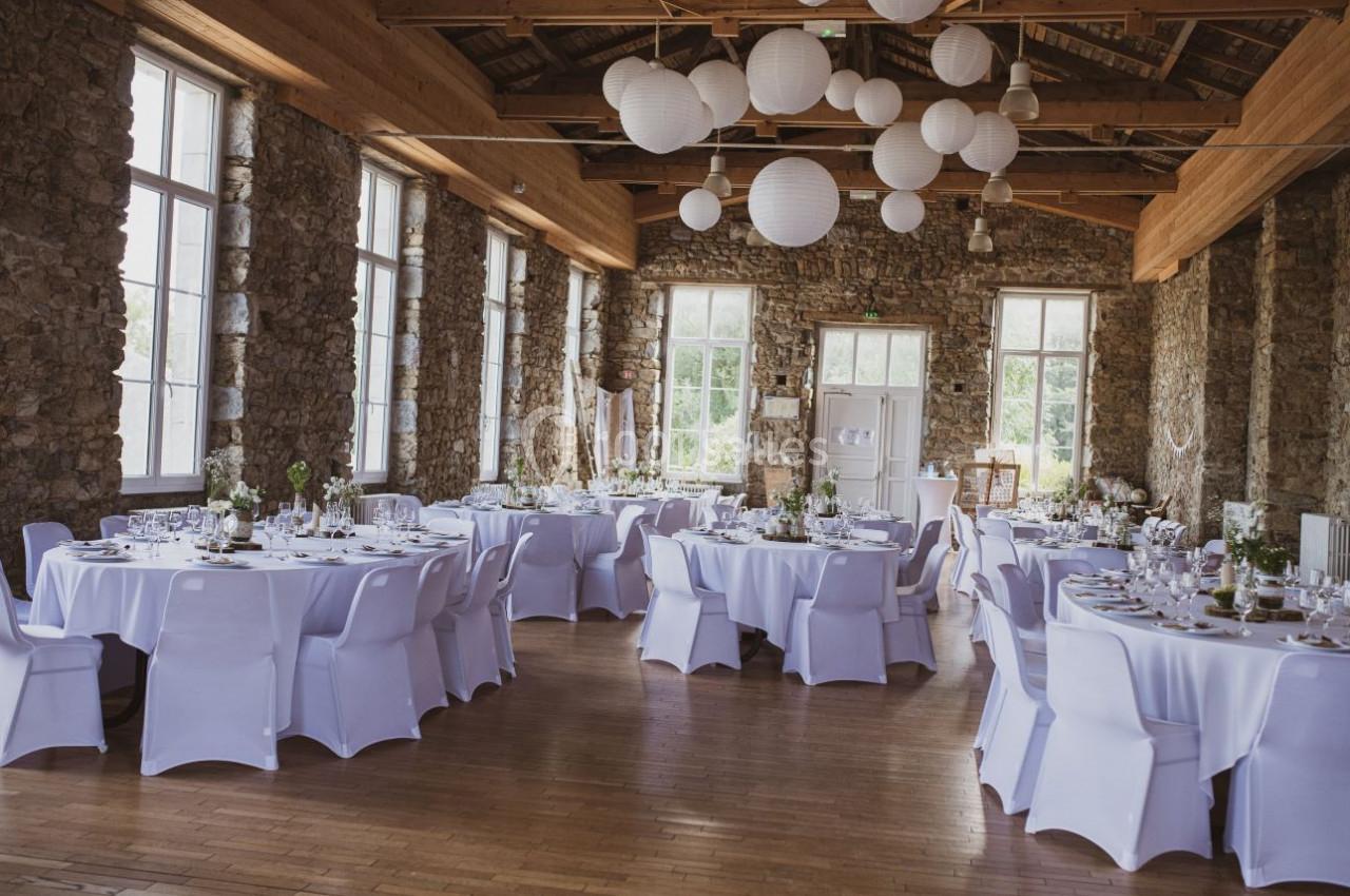 Salle de réception lumineuse avec murs en pierre, tables rondes dressées et décorations suspendues en papier blanc.