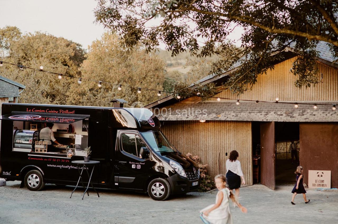 Camion de cuisine stationné devant une grange en bois, avec des enfants jouant à proximité sous des guirlandes lumineuses.
