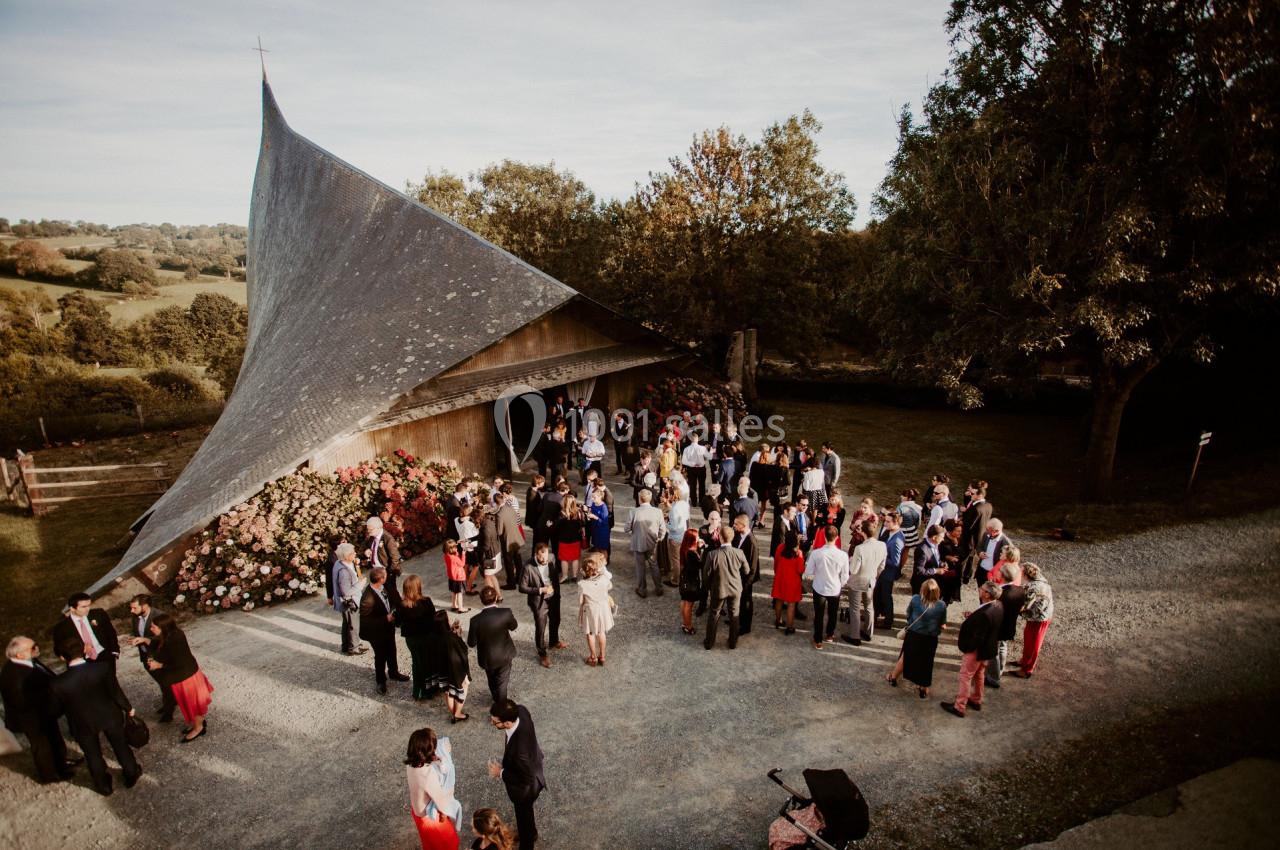 Groupe de personnes rassemblées devant une petite chapelle en bois entourée de végétation, par une journée ensoleillée.