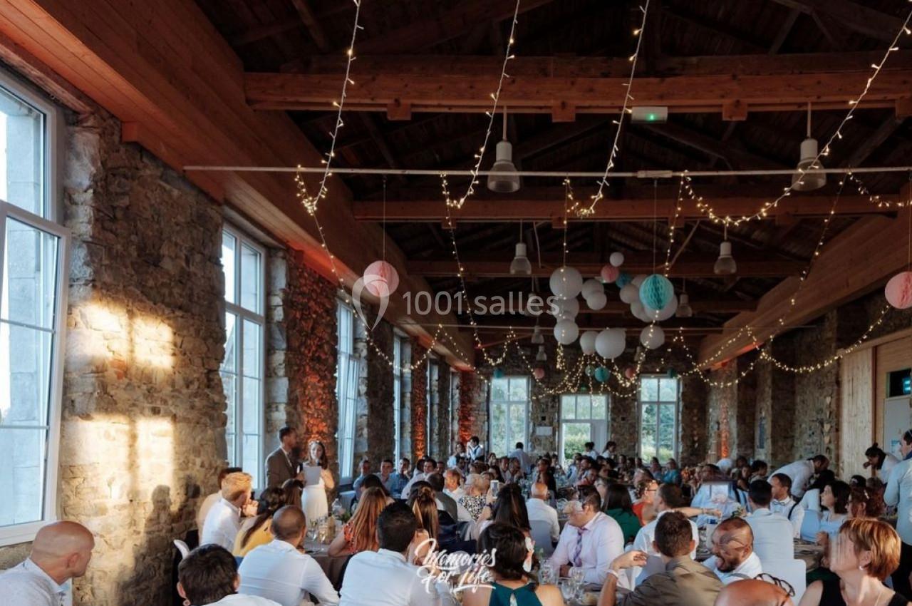 Salle en pierre avec de grandes fenêtres, décorée de guirlandes lumineuses, accueillant un dîner avec de nombreux convives.