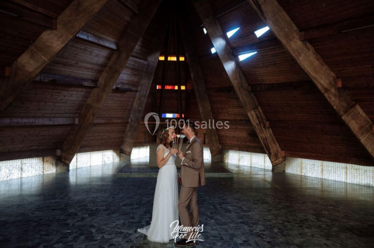 Un couple en tenue de mariage danse dans une salle en bois avec des vitraux colorés et un sol en pierre.